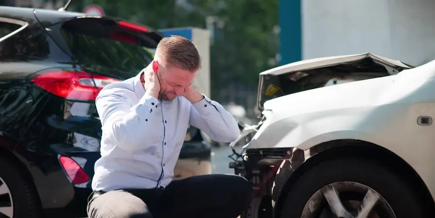 A distressed man in a white shirt is holding his neck in pain and is sitting beside damaged cars after a neck injury accident collision in Atlanta.
