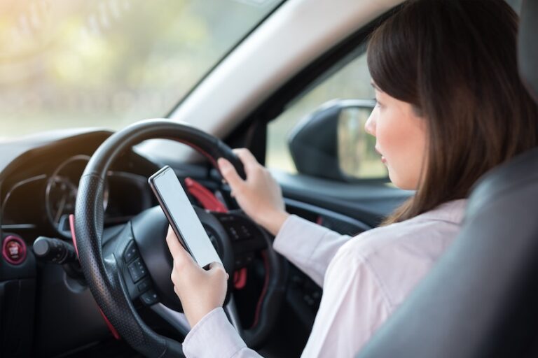 A female driver holds a smartphone in one hand while gripping the steering wheel with the other while driving illustrating the type of distracted driving behavior behind the wheel of a rideshare vehicle that commonly causes a Lyft accident in Metro Atlanta.