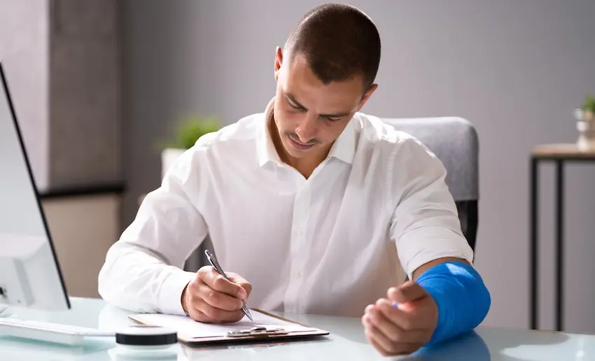 A young man with a blue arm cast writes on a clipboard at an office desk beside a computer representing a rideshare passenger documenting injuries sustained in an Uber accident in order to file a personal injury claim with help from Atlanta Metro Law.