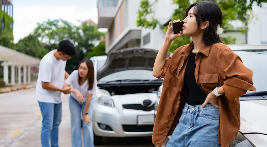 A young woman leans against the hood of a damaged car with its hood raised while talking on her phone as two other people inspect an injury behind her depicting the chaotic aftermath of an Uber accident where passengers and drivers must immediately report the collision in Atlanta.