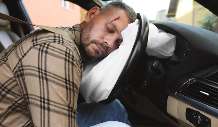 A bearded man in a plaid shirt is slumped unconscious over a deployed white airbag with a bleeding gash on his forehead showing the severe and life-threatening injuries a driver can sustain in a head-on collision in Metro Atlanta.