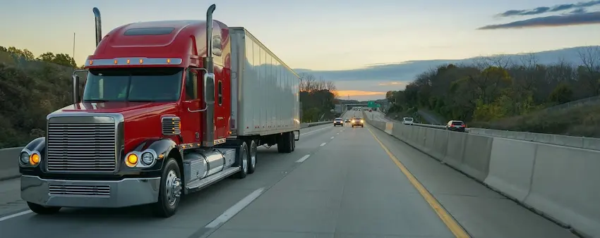 A red semi-truck with a large silver cargo trailer dominates the center lane of a multi-lane highway at dusk with other vehicles visible ahead and tree-lined hills and a colorful sky in the background depicting the type of large commercial vehicle that shares the road with passenger cars and poses significant injury risks in the event of a crash in Metro Atlanta.