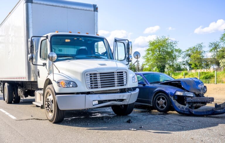 A large white commercial vehicle with a box trailer sits stopped on a sunny road with its front end pushed against a badly damaged blue sedan whose bumper and hood have been completely torn away and left scattered on the pavement illustrating the catastrophic destruction a commercial vehicle can inflict on a passenger car in a collision in Metro Atlanta.