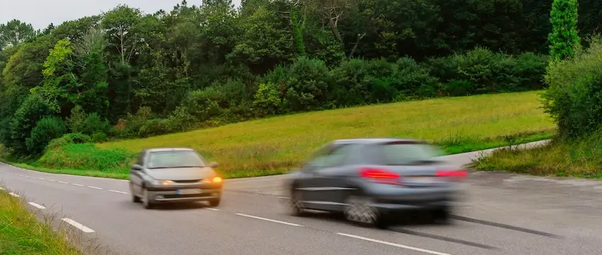 Two sedans traveling in opposite directions on a two-lane rural road lined with dense green trees and open fields with one car crossing the center line toward oncoming traffic depicting the dangerous split-second moment that can lead to a devastating head-on collision and could use help from Atlanta Metro Law.